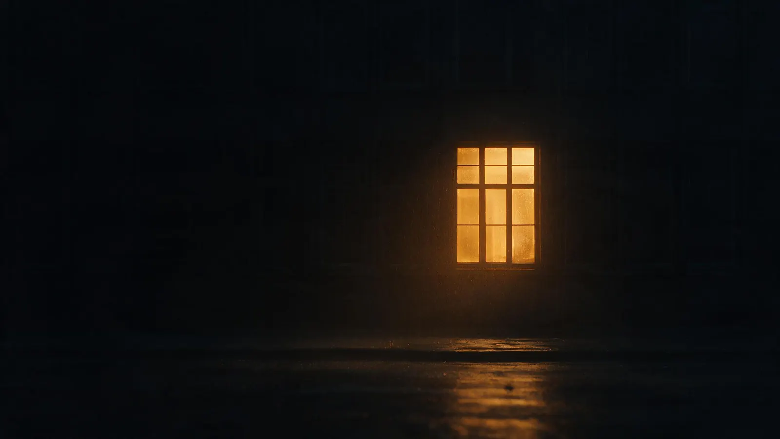 A single window lit warm amber in a wall of dark windows at night, its light reflected on wet ground.