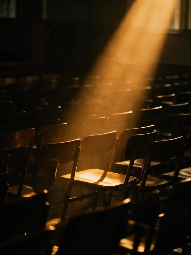Rows of empty classroom chairs in a dark hall, one shaft of light across a single chair.